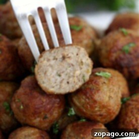 A fork holds a halved meatball over a pile of oven-cooked meatballs, perfectly garnished with herbs.