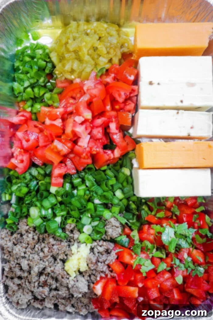 Meaty Smoked Queso Dip 5 Prepped ingredients for smoked queso dip, featuring chopped vegetables, various cheese blocks, and cooked ground meat arranged in a foil container for assembly.