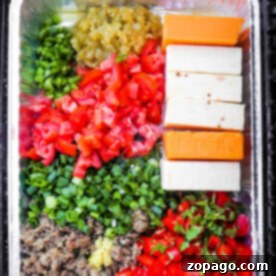A tray with diced vegetables, cheese blocks, and cooked meat in a foil container on the grill melds into a savory smoked queso dip.