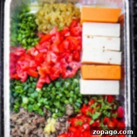 A tray with diced vegetables, cheese blocks, and cooked meat in a foil container on the grill melds into a savory smoked queso dip.
