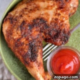 A cooked chicken leg is served on a green plate with a small glass bowl of ketchup, offering inspiration for those wondering how to cook turkey legs and thighs. A fork rests beneath the succulent meat, and a small bowl with a white substance peeks from the bottom left corner.