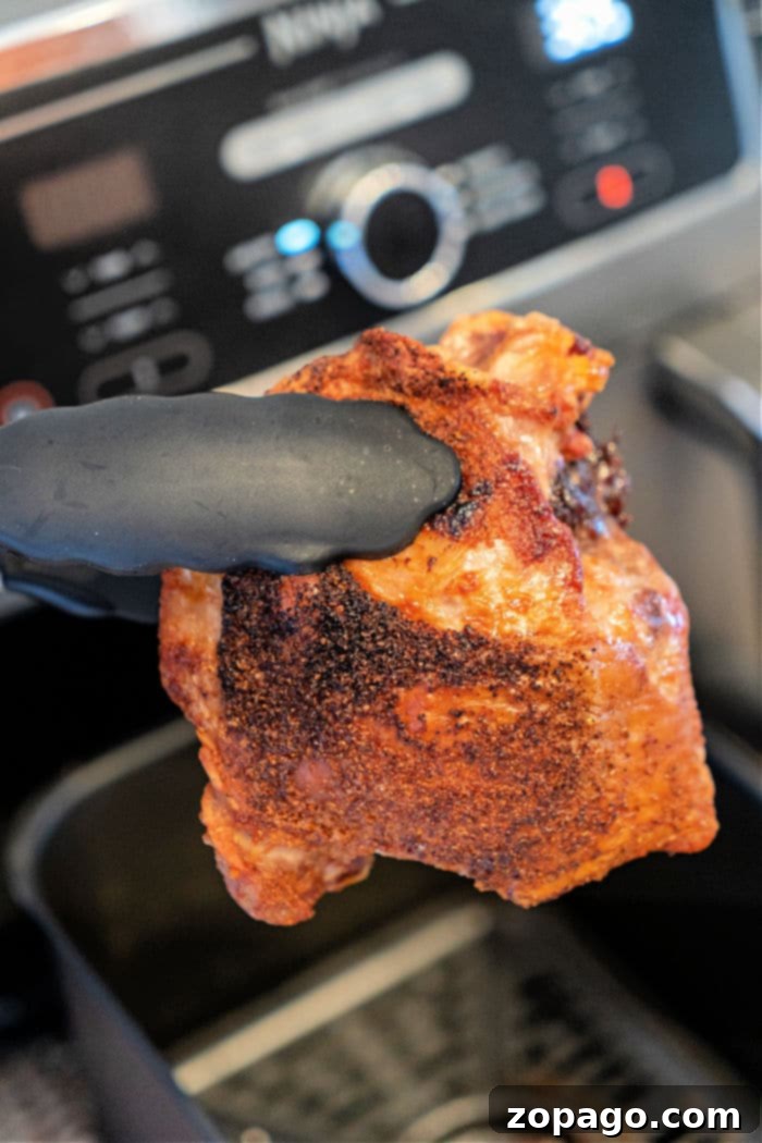 Succulent Turkey Leg Quarters 7 A gloved hand holds a piece of cooked, seasoned turkey thigh in front of an air fryer. The meat boasts a crispy, browned exterior. In the background, the air fryer display and control knobs stand ready for your next culinary adventure.