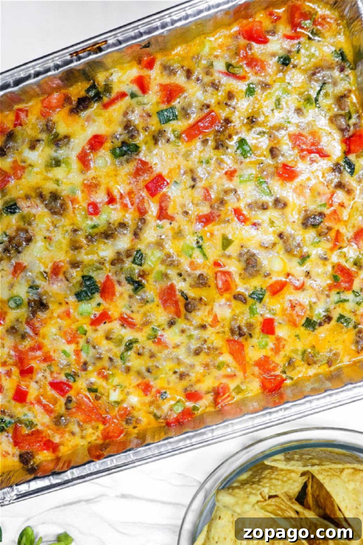 Baked breakfast casserole with sausage, cheese, colorful vegetables, and a hearty queso dip with meat in a foil tray, served next to tortilla chips.