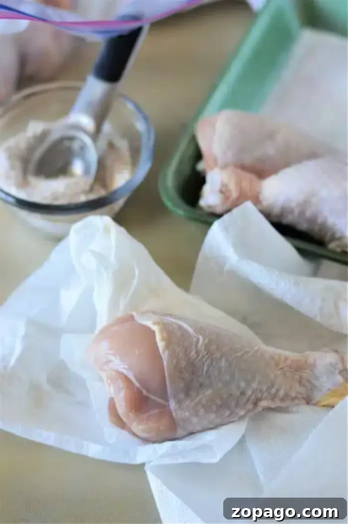 Crispy Baked Chicken Legs 3 Raw chicken drumsticks are on white paper towels, ready for preparation. A small scoop rests in a bowl nearby containing a flour mixture. A green tray in the background holds additional chicken drumsticks, soon to become baked chicken legs.