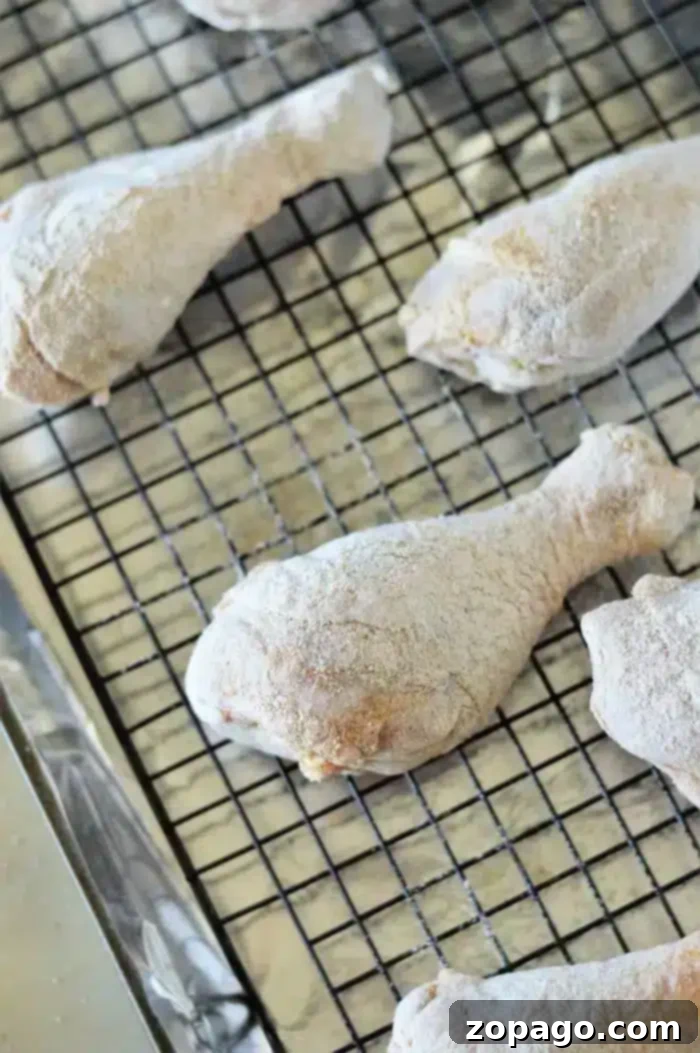 Crispy Baked Chicken Legs 5 Baked chicken legs are coated in flour and placed on a wire rack for drying. The rack sits on a baking sheet lined with foil, suggesting the drumsticks are being prepared for frying or baking.