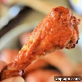 A hand holding a crispy, saucy baked chicken leg, reminiscent of buffalo chicken drumsticks, with a blurred background.