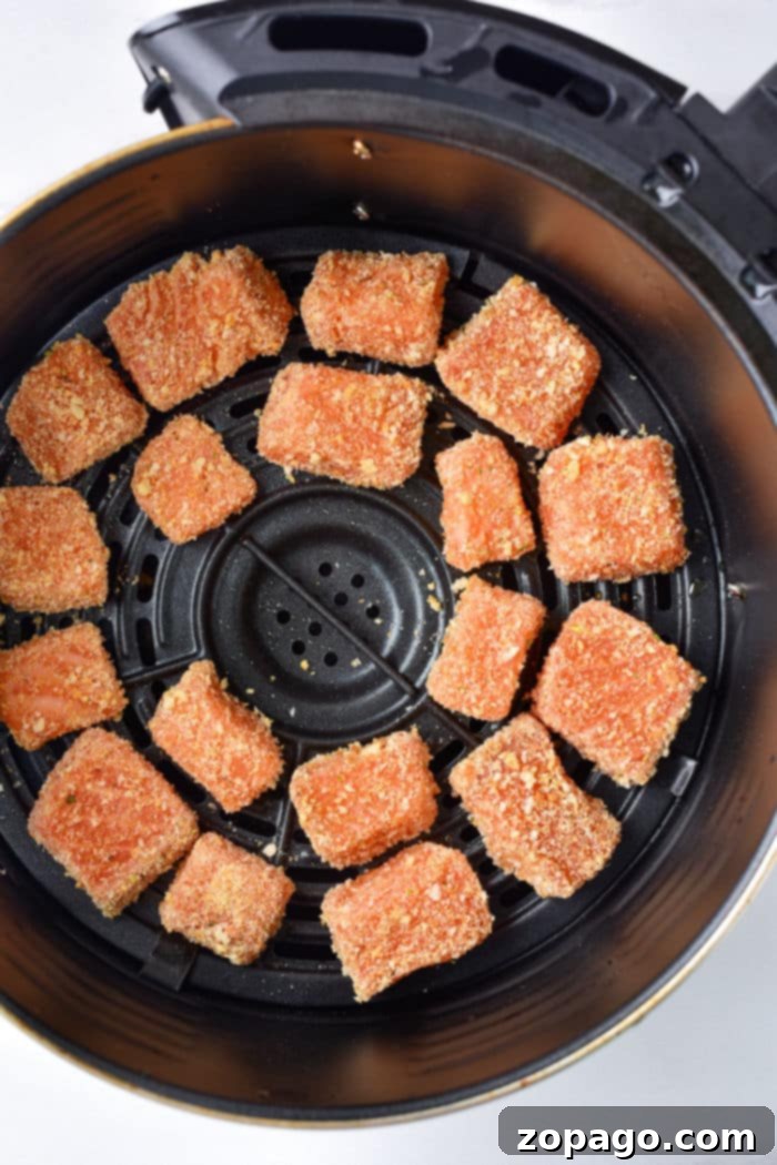 Breaded salmon pieces, similar to delectable salmon bites, sit in an air fryer basket, ready to be cooked to a perfect crisp.