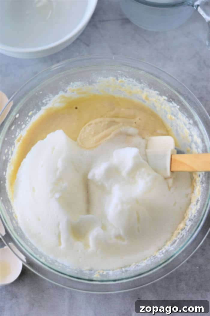 No Baking Powder Pancakes 3 A glass bowl containing whipped egg whites being gently folded in using a spatula. The bowl rests on a gray countertop, with another bowl and kitchen items visible in the background.