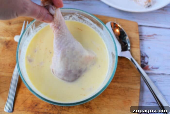 Pan Fried Chicken Legs with Flour 4 Hand dipping raw chicken legs into creamy batter in a glass bowl on a wooden board, ready to be transformed into delicious pan fried chicken legs, with a spoon and fork beside it.