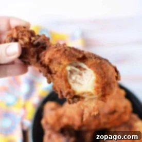 A close-up captures a hand holding a fried chicken drumstick with a bite taken out, hovering over a bowl full of crispy fried chicken legs.
