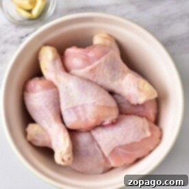 Raw chicken drumsticks in a bowl on a marble surface, accompanied by garlic cloves and a bowl of mayonnaise nearby, ready to inspire the best chicken leg recipes for your next culinary adventure.