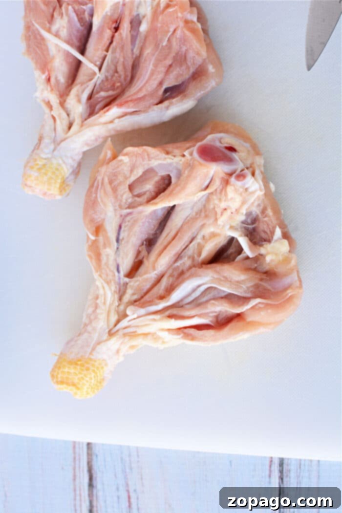 Crispy Oven Butterfly Chicken 4 Two raw, butterflied chicken legs rest on a white cutting board, partially cut open, with a knife visible in the top right corner, ready for seasoning.