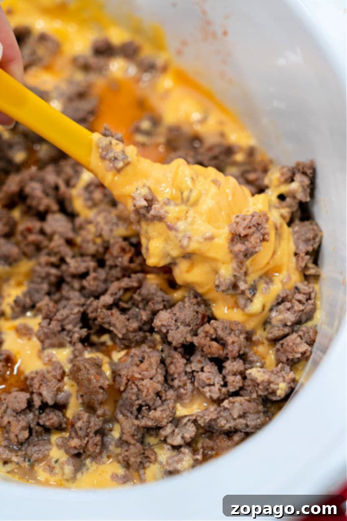 Melted Velveeta cheese and browned ground spicy sausage are being stirred in a slow cooker with a yellow spatula, creating a delicious Sausage Dip.