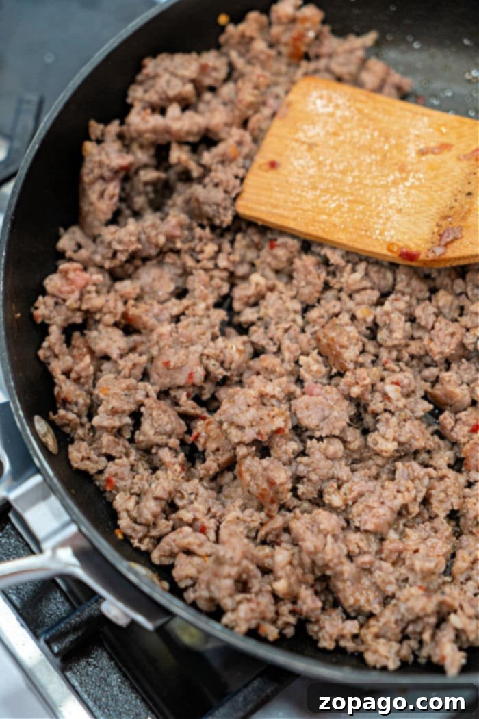 Ground sausage sizzles in a black skillet on the stovetop, stirred gently with a wooden spatula, creating a perfect base for a hearty Velveeta and Rotel dip.