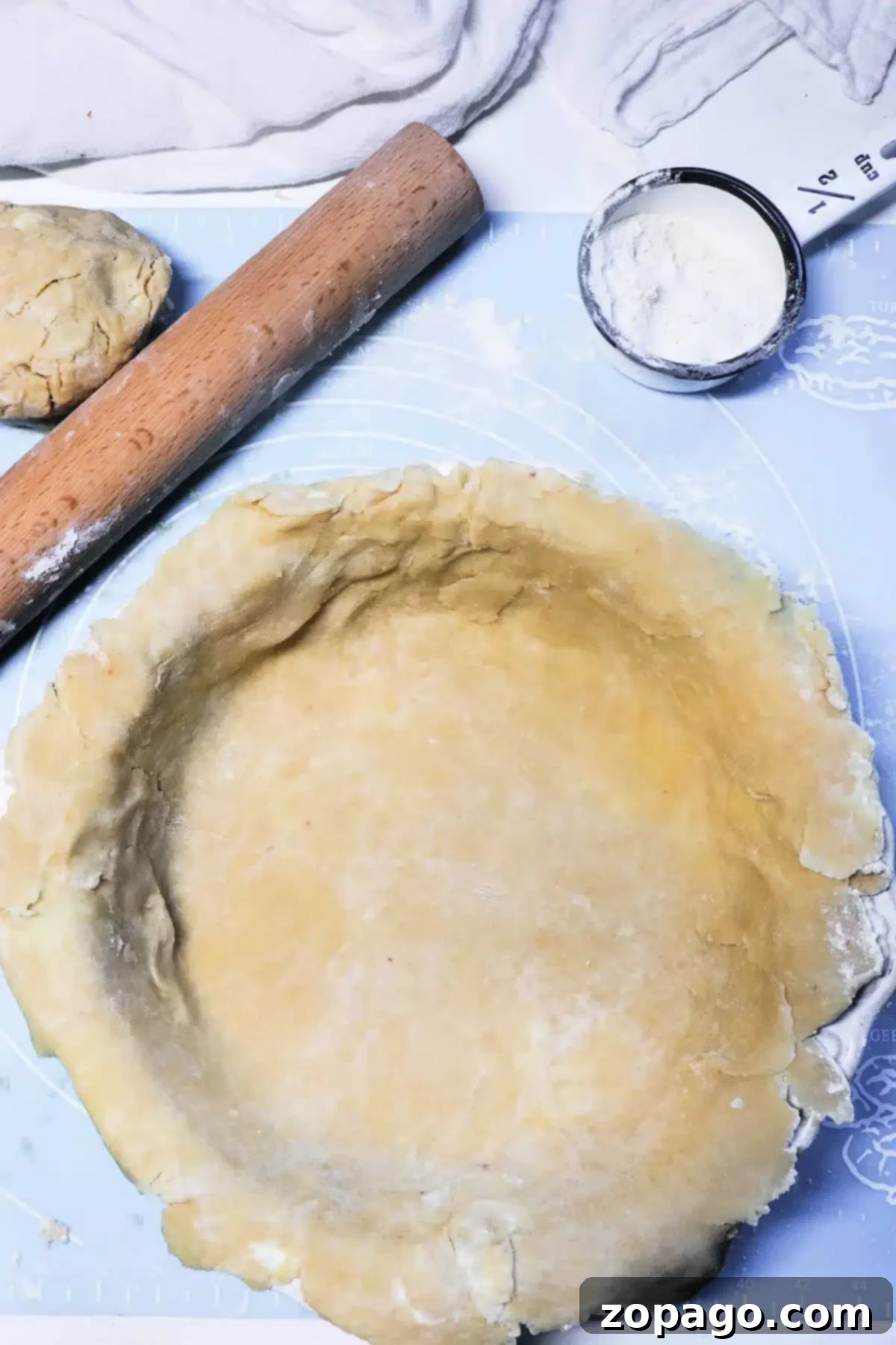 Buttery Pot Pie Crown 2 A rolled-out pot pie crust is placed in a pie dish on a blue work surface. Nearby are a wooden rolling pin, a ball of dough, and a tin measuring cup with flour. White fabric is in the background.