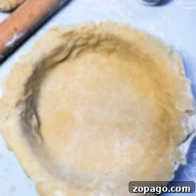 Flaky Pot Pie Lid 8 A rolled-out pot pie crust is placed in a round pie dish on a light blue surface. A wooden rolling pin and a small cup of flour are visible nearby, ready for adjustments. The dough has a slightly uneven edge, adding to its homemade charm.