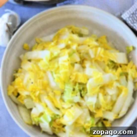 A bowl of cooked napa cabbage sits on the table, accompanied by a pan and a hint of garlic in the background, offering a delightful glimpse into a simple yet flavorful recipe.