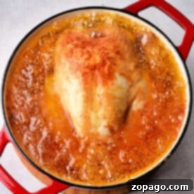 Whole chicken simmering in bubbling broth inside a red Dutch oven on a grey surface, ready to become the juiciest Whole Fried Chicken.