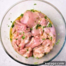 Raw, marinated chicken pieces, infused with a Mediterranean chicken marinade and herbs, rest in a clear glass bowl on a light surface.