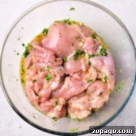 Raw, marinated chicken pieces, infused with a Mediterranean chicken marinade and herbs, rest in a clear glass bowl on a light surface.