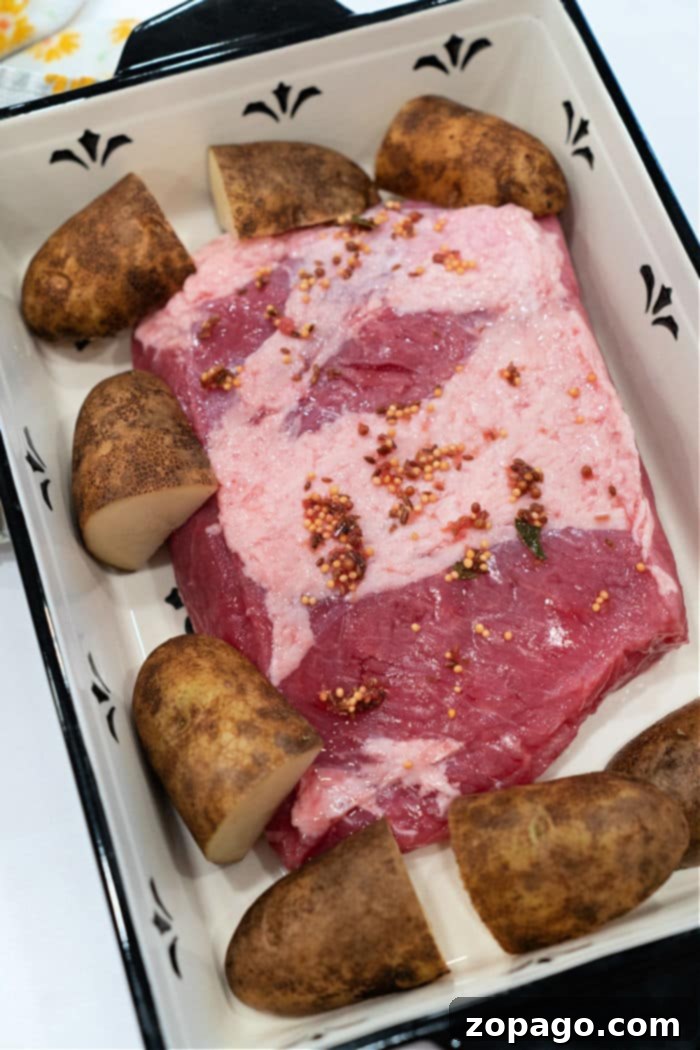 Perfect Oven Corned Beef 4 A raw, seasoned corned beef brisket positioned in a rectangular baking dish, surrounded by halved potatoes. The dish is a simple white with black trim, ready for the oven to begin cooking this classic meal.