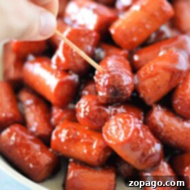 A hand holding a toothpick picks up a glazed mini sausage from a bowl of hot dog burnt ends.