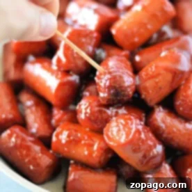 A hand holds a toothpick, picking up a glazed mini sausage from a bowl of hot dog burnt ends.