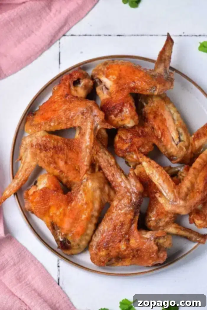Crispy Baked Whole Chicken Wings 2 A plate of baked whole chicken wings is displayed on a white surface with a pink cloth and a few green herbs scattered around. The wings are golden-brown and appear crispy.