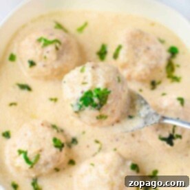 Creamy Alfredo Meatballs 5 A bowl of creamy white sauce with meatballs, garnished with chopped herbs. A spoon lifts one Alfredo meatball, enticing you to savor its rich flavor.