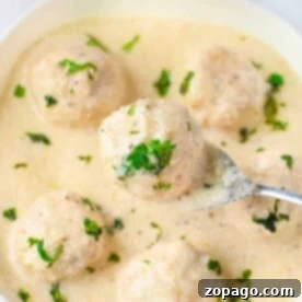 A bowl of creamy white sauce with meatballs, garnished with chopped herbs. A spoon lifts one Alfredo meatball, enticing you to savor its rich flavor.
