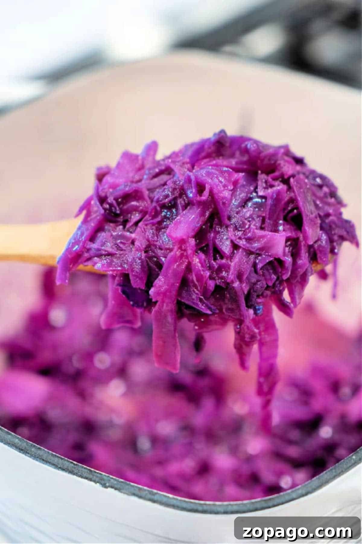 Sweet and Sour Red Cabbage 2 A wooden spoon holding a portion of vibrant sautéed red cabbage over a pot filled with more shredded cabbage, ready to be served.
