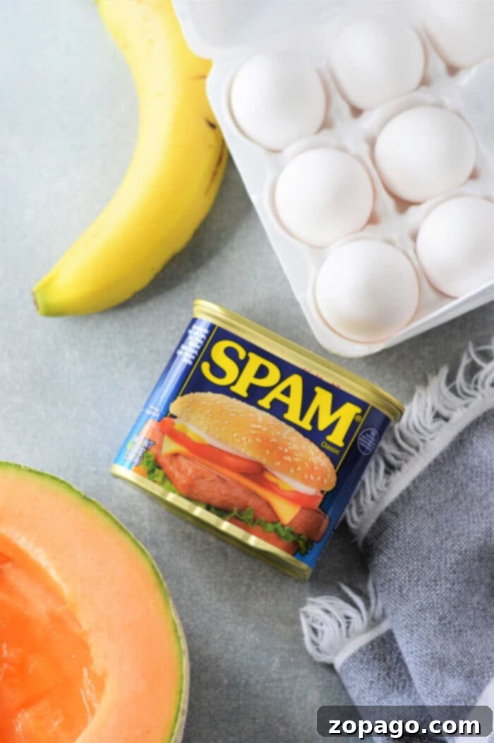 A can of Spam, a carton of eggs ready for a delicious Spam and Eggs recipe, a ripe banana, half a cantaloupe, and a gray cloth rest on the countertop.