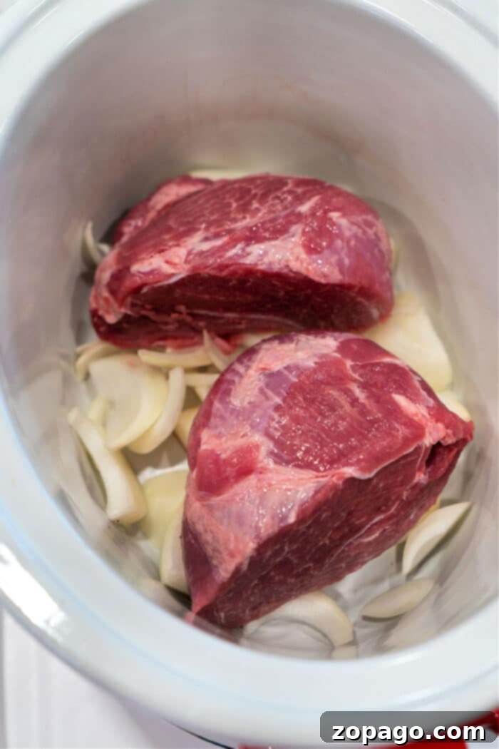 Two raw pork picnic roast rest in a slow cooker, surrounded by sliced onions.