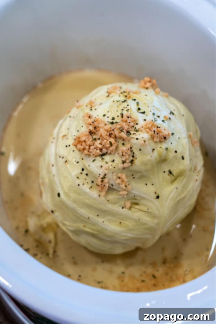 Slow Cooked Whole Cabbage Perfection 5 A whole head of cabbage, slowly cooked to perfection in a white dish, is beautifully topped with minced garlic and herbs nestled in a savory broth.