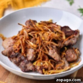 A plate of grilled chicken liver topped with crispy fried onions sits on the table, tempting you to pick up the nearby fork. This dish perfectly complements a guide on how to cook chicken liver to perfection.