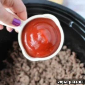 A hand holds a measuring cup of ketchup, poised above a pot of cooked ground meat—the beginning steps for crafting delicious slow cooker sloppy joes.
