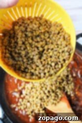 A hand demonstrating how to cook green lentils on the stove, pouring them from a yellow strainer into a pot of sauce with a wooden spoon.