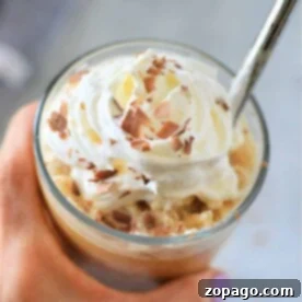 Coffee Frappe 9 Close-up of a hand holding a frozen iced coffee topped with whipped cream and chocolate shavings, with a spoon.
