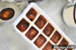 Blended Iced Coffee 4 A white ice cube tray filled with brown liquid, presumably frozen coffee, is placed on a light gray countertop. To the left of the tray, a teaspoon holds a small amount of the same brown liquid. A gray cloth and a glass pitcher are partially visible in the background, hinting at how to make cold foam for coffee.