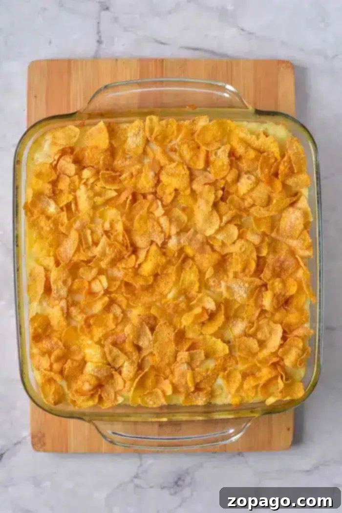 Easy 5 Ingredient Hash Brown Casserole 2 A glass baking dish filled with a golden 5-Ingredient Hash Brown Casserole topped with crispy cornflakes, placed on a wooden cutting board on a marble countertop. The casserole looks creamy and inviting beneath its crunchy crust.