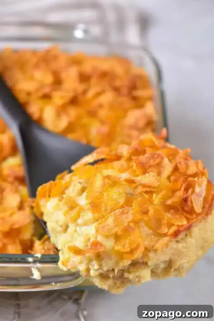 Easy 5 Ingredient Hash Brown Casserole 6 A close-up shot of a serving of cheesy 5 Ingredient Hash Brown Casserole, topped with golden, crispy cornflakes, being lifted from a glass baking dish with a black serving spoon. The background is softly blurred.
