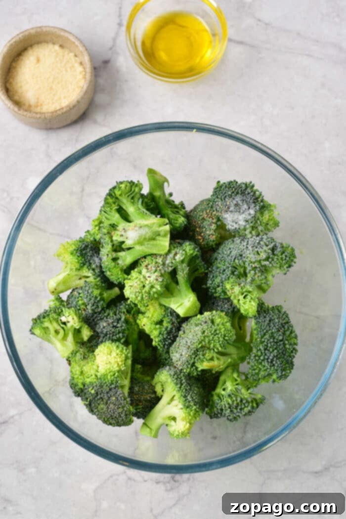 Perfectly Baked Frozen Broccoli 3 A glass bowl with frozen broccoli florets, perfect for baking, sits next to small bowls of grated cheese and olive oil on a marble surface, highlighting the simple ingredients needed.