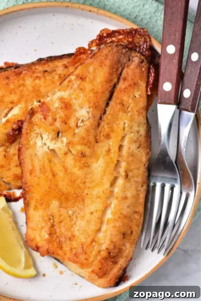 Grilled fish fillets with a golden crust rest on a white plate, alongside a lemon wedge. Perfectly cooked, just like baking frozen fish to perfection. A set of silverware with wooden handles is placed beside them, with a green textured cloth partially visible in the background.