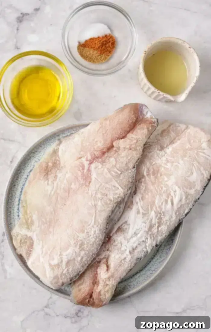 Deliciously Baked Frozen Fish 3 Two raw fish fillets sit on a plate, surrounded by small bowls containing olive oil, seasoning mix, and a light-colored liquid on a marble surface—ideal ingredients when baking frozen fish to perfection.