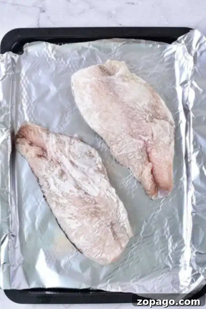 Deliciously Baked Frozen Fish 4 Two raw fish fillets rest on a baking sheet lined with aluminum foil, ready for baking. Their light, frosty appearance suggests they're frozen. The simple, light background enhances their fresh allure.