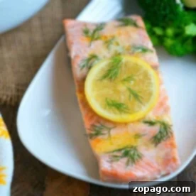 Salmon fillet garnished with a lemon slice and dill on a white plate, showcasing one of our delightful salmon dinner recipes, accompanied by broccoli in the background.