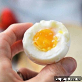 A hand holding a half-peeled soft-boiled egg reveals a perfectly runny yolk, the result of knowing just how long to cook soft boiled eggs on the stove.
