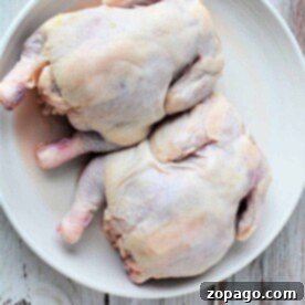Two whole uncooked cornish hens rest on a white plate against a rustic wooden background, inviting the question: how long to cook Cornish hens to achieve that perfect balance of tenderness and flavor?.