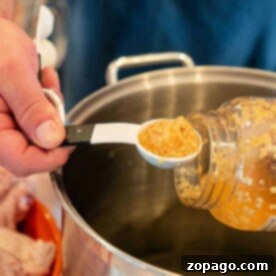 A person holds a jar of minced garlic, using a white measuring spoon to scoop it into a large metal pot for their whole chicken brine. A plastic bag with uncooked chicken wings is visible in the background.