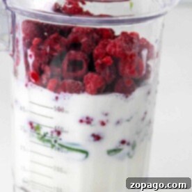A blender jar filled with milk, frozen raspberries, and greens is prepped and ready to show you how to make a thick smoothie.